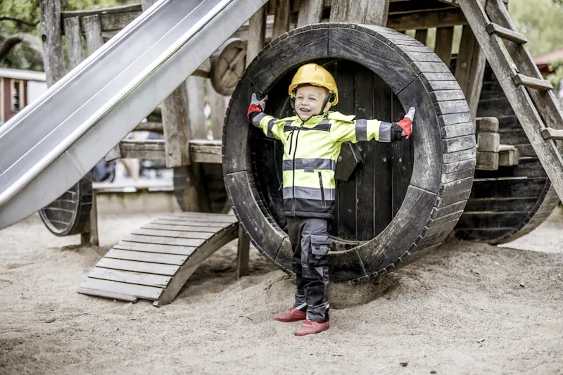 Kind mit gelbem Schutzhelm, reflektierender Arbeitsjacke, Arbeitshose und roten Schuhen steht vor großem Holzrad auf Sandboden und breitet die Arme aus